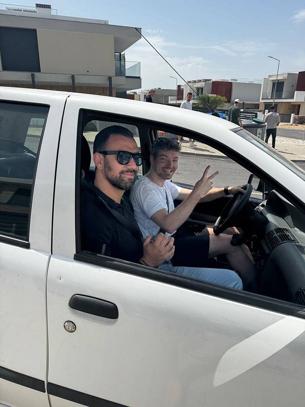 Two men are sitting in a white car, smiling and posing for the camera.