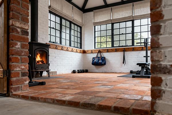 A cozy farmhouse-style home gym with terracotta brick paver flooring and white-painted brick walls. A black cast-iron wood-burning stove glows beside an exposed brick column. Black steel-framed windows with textured glass and light linen roman shades wrap two walls above a continuous oak peg rail holding a blue duffel and a training strap. A bench and free weights sit on a rubber mat, with extra dumbbells in the corner and dark rafters overhead.