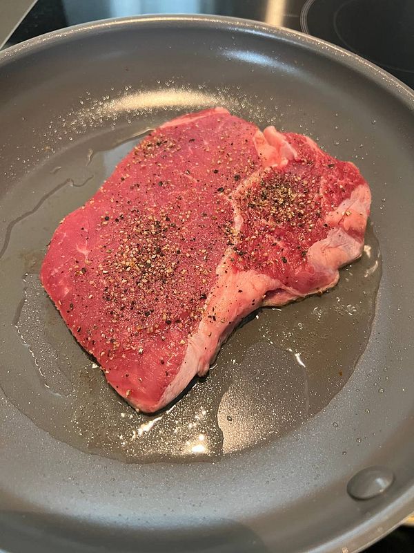 A raw steak seasoned with black pepper is placed in a frying pan.