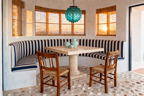 A curved built-in banquette upholstered in navy-and-white stripes wraps a small dining alcove around a square travertine pedestal table. Two wooden chairs with woven rush seats face the table, which holds seafoam ceramic vessels. A turquoise bubble-glass pendant hangs above, while three windows with woven bamboo shades filter warm sunset light. Textured plaster walls and a concrete floor inlaid with seashells complete the breezy coastal setting.