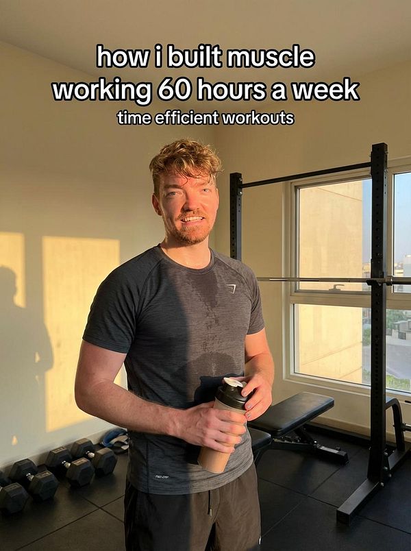 A fit individual stands in a gym, holding a protein shake and smiling, with workout equipment in the background.