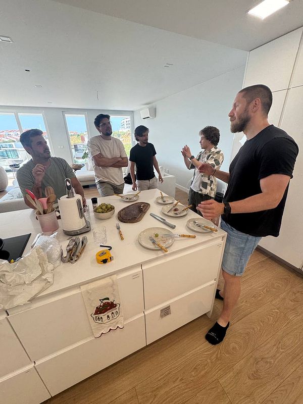 A group of five men are engaged in conversation around a kitchen island with plates and utensils.
