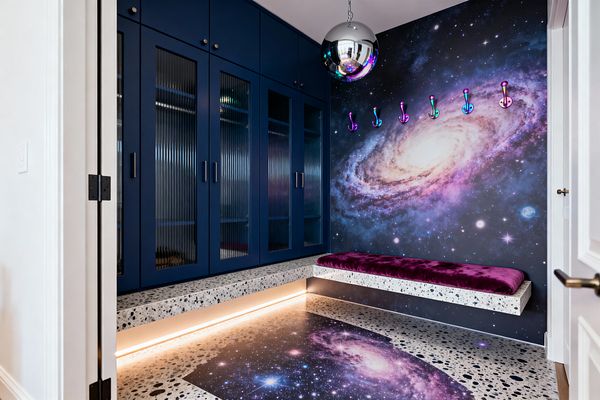 A mudroom with deep navy built-in cabinets featuring vertical reeded-glass doors faces a wall-sized galaxy mural that continues as a matching inlay on a speckled terrazzo floor. A floating terrazzo bench topped with a plush magenta velvet cushion sits beneath iridescent hooks, warm LED strip lighting glows under the bench and toe kick, and a polished chrome globe pendant reflects the saturated purples, blues, and starry imagery across the space.