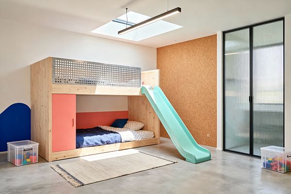 A bright modern kids’ room with a light pine bunk bed featuring a perforated metal safety rail and a mint-green slide, set on a polished concrete floor. Coral sliding panel and headboard, a blue wave wall accent, a full cork accent wall, ribbed glass sliding doors with black frames, a skylight with a linear pendant, a flat-weave rug, and clear plastic toy bins complete the minimalist, playful space.