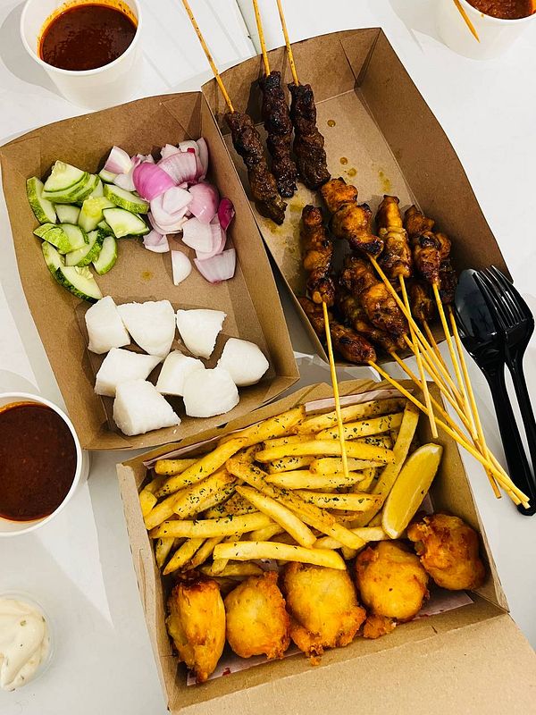 Overhead view of a wooden platter with satay skewers, rice cake, cucumber slices, onion pieces, and a side of sauce.