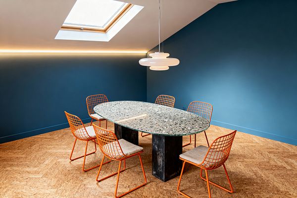 A modern dining room features deep teal walls and a sloped ceiling with a skylight and a slim LED strip. At center sits an oval terrazzo table on two blackened metal pedestals, encircled by six orange powder-coated wire chairs with light beige cushions, set on a warm herringbone cork floor, and lit by a white opal-glass pendant.