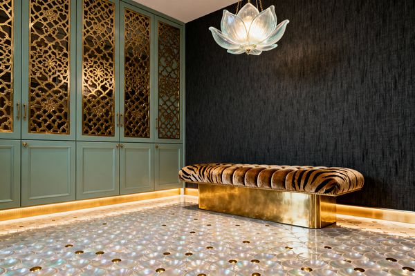 A glamorous mudroom showcases pale sage built-in cabinets with mirrored doors overlaid in ornate brass fretwork and polished pulls, all lit by warm toe-kick LEDs. A charcoal grasscloth wall frames a channel-tufted tiger-stripe velvet bench on a brass plinth, while a lotus-shaped blown-glass pendant glows above and an iridescent mother-of-pearl scallop floor punctuated with small brass dots reflects the golden light.