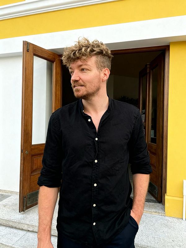 A man with curly hair stands outside a building with a yellow wall and wooden doors.