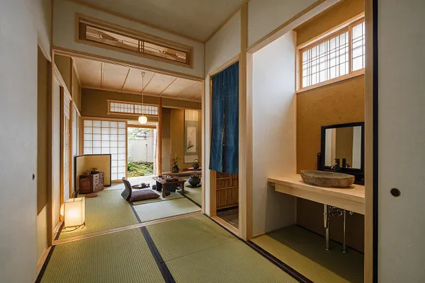 A Japanese Zen-style powder room with tatami mats, light wood trim, and a simple stone vessel sink on a pale wood vanity. Shoji-style screens, a blue fabric curtain, and soft natural light create a calm, minimalist atmosphere.