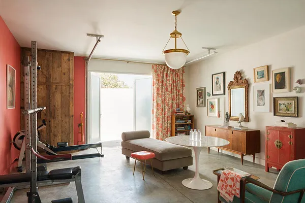 A bright eclectic home gym combines workout equipment with mid-century and vintage furnishings. The room has coral and white walls, a concrete floor, a rustic wood door, floral curtains, framed artwork, a gold-and-white pendant light, and a mix of a chaise lounge, marble pedestal table, and colorful storage pieces.