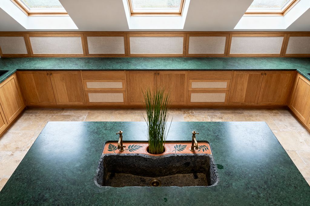 A bright kitchen with U-shaped oak cabinetry and deep green stone countertops sits beneath three large skylights. The island features a rough stone trough sink flanked by brass taps, set into a terracotta deck stamped with leaf motifs and a circular planter holding tall grass-like greenery. Light beige textured wall panels run above the counters, and beige limestone floor tiles complete the warm, natural look.
