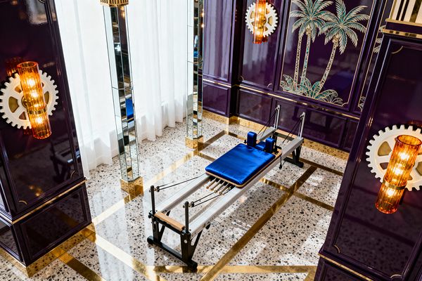 Luxurious home gym featuring a Pilates reformer with aluminum rails, black supports, and a royal-blue upholstered carriage sits on a terrazzo floor patterned with polished brass bands. Surrounding walls are high-gloss purple panels with gold trim, palm tree artwork, and amber ribbed sconces, while mirrored columns and sheer white curtains flood the space with reflected light, creating a glamorous Hollywood Regency atmosphere.