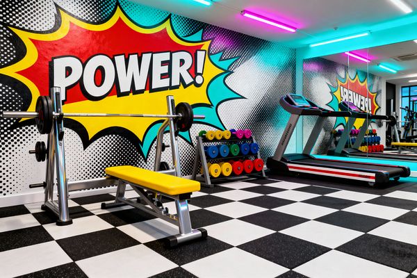 Bright Pop Art home gym with a large comic-style mural reading 'POWER!' in a yellow, red, and cyan starburst over a halftone dot background. The room features a chrome weight bench with a yellow vinyl seat, a rack of multicolored dumbbells, a striped treadmill facing a mirrored wall, black-and-white checkered flooring, and ceiling-mounted magenta and aqua light bars that bathe the space in bold color.