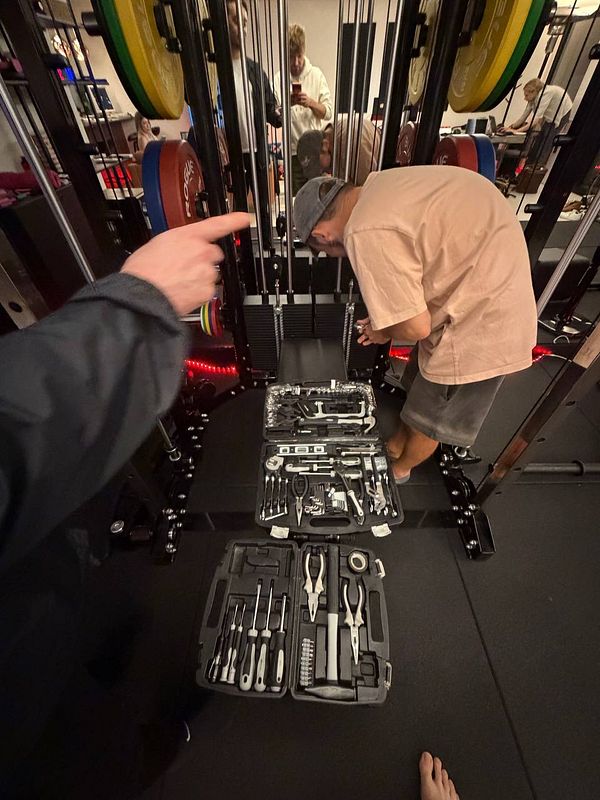 A person working with tools on a gym machine while another person points toward the area, with a reflection showing a third person taking the photo.