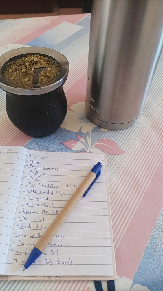 A chic workspace setup featuring a laptop with an artistic wallpaper, a MacBook, a cup of yerba mate tea, notebooks, and stationery on a white desk.
