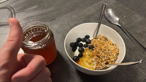A hand gives a thumbs up next to a jar of honey and a bowl of yogurt topped with granola and blueberries.