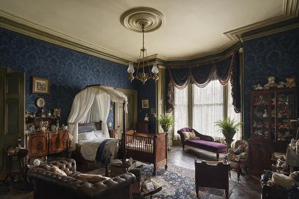 A richly decorated Victorian nursery with dark blue patterned wallpaper, a carved wooden cradle bed under a white canopy, and heavy draped window treatments. The room includes antique wooden cabinets, a tufted leather sofa, upholstered seating, and vintage decor arranged around a large patterned area rug.