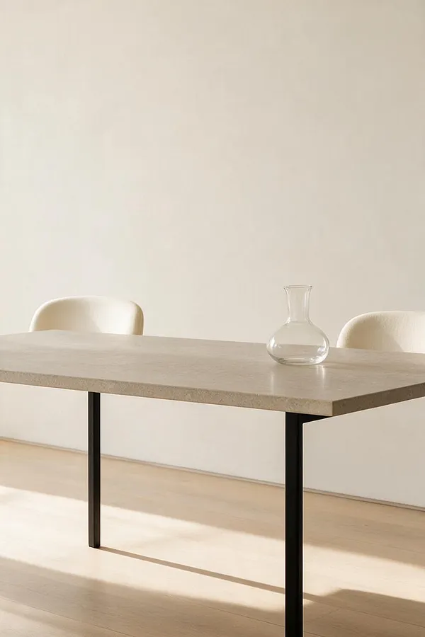 A sparse minimalist dining vignette with a pale stone or concrete table, slim black metal legs, and two cream upholstered chairs partially visible on either side. A small clear glass carafe sits on the tabletop against a plain off-white wall and light wood floor.