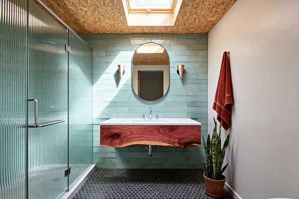 A mid-century modern bathroom with a fluted-glass shower on the left, a wall of mint-green subway tiles, and a floating vanity faced in a live-edge solid wood slab topped by a light gray countertop. An oval frameless mirror and two cone-shaped brass sconces frame the sink, while a charcoal hex tile floor, OSB ceiling with a skylight, rust towel, and a potted snake plant complete the space.