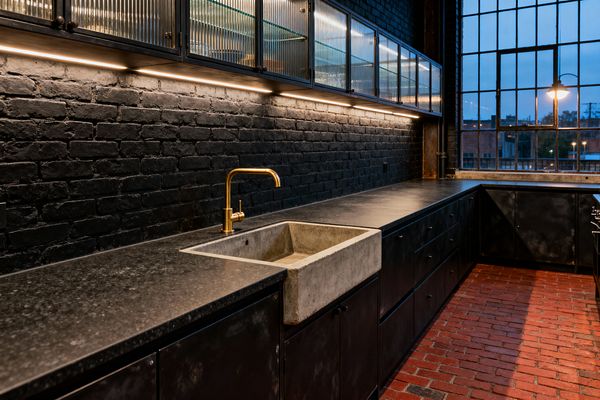 A moody industrial kitchen features matte black cabinets and a long dark stone countertop against a black painted brick wall. A concrete apron-front sink is paired with a warm brass gooseneck faucet. Upper cabinets with metal frames and fluted glass sit above continuous LED under-cabinet lighting. The floor is red brick, and a large factory-style grid window brings evening light, alongside an exposed sconce and piping for an authentic loft feel.