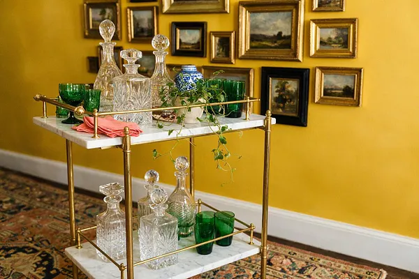 A brass bar cart with white marble shelves is styled with cut-crystal decanters, green glassware, a small potted vine, and a folded pink napkin. It sits in front of a mustard-yellow wall filled with a clustered gallery of small framed landscape paintings and above a patterned rug.
