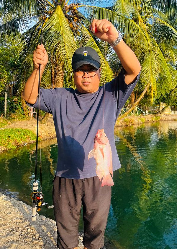 A young man holding a caught fish near a body of water with a city skyline in the background.