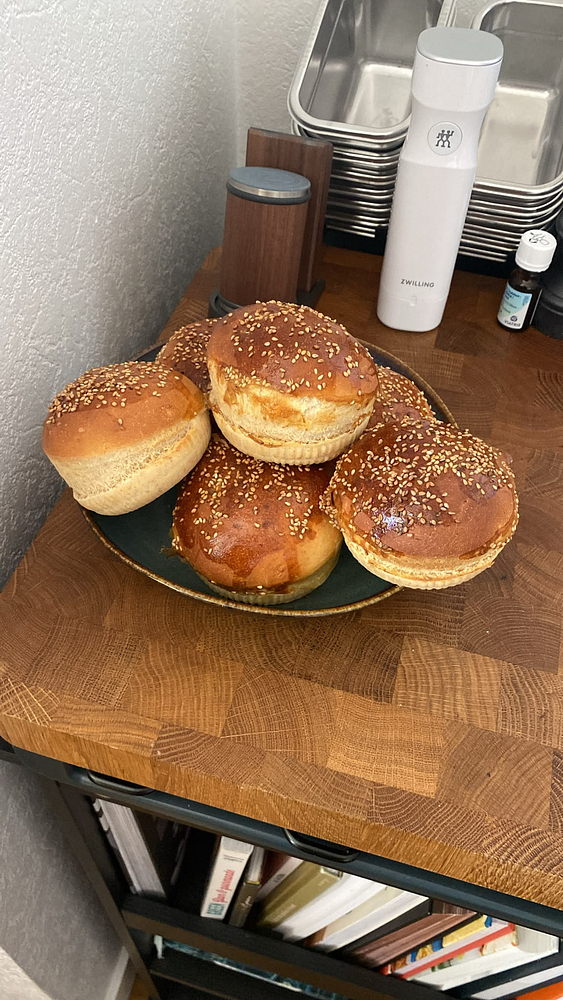 A plate of freshly baked sesame seed buns on a wooden countertop with kitchen items in the background.