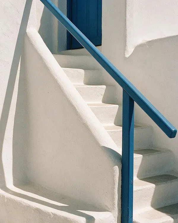 A Mediterranean-style exterior staircase with smooth white stucco walls and built-in steps, accented by a vivid cobalt blue handrail and matching door shutters. Strong sunlight creates crisp shadows across the curving surfaces, emphasizing the simple, sculptural architecture.