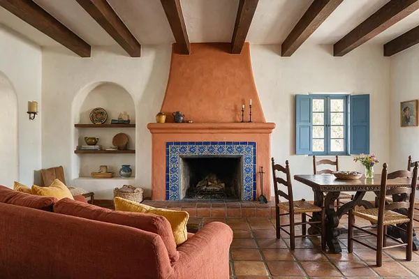 A Mediterranean-style living room with white stucco walls, exposed dark wood ceiling beams, and a large terracotta plaster fireplace trimmed in blue patterned tiles. The room includes a rust-colored sofa, a carved wood dining table with woven-seat chairs, terracotta floor tiles, and a blue-shuttered window that adds soft natural light.