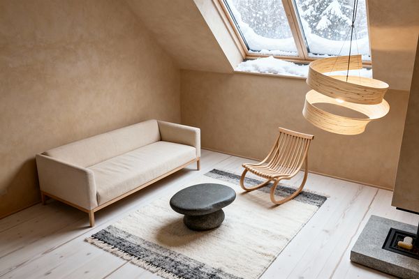 A minimalist Scandinavian loft living room beneath a sloped ceiling shows a light linen sofa on a slim wood base, a curved slatted wooden lounge chair, and a pebble-shaped dark stone coffee table atop a cream wool rug with charcoal borders. Warm beige limewashed walls, whitewashed pine floorboards, a layered wood-veneer pendant, and large skylight windows with snow outside create a calm, natural atmosphere beside a low stone hearth and modern firebox.