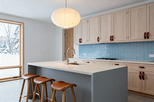 Bright Scandinavian kitchen with a large gray waterfall island topped in light beige stone and a stainless gooseneck faucet. Along the back wall, light oak Shaker cabinets with cognac leather strap pulls sit above a sky-blue square tile backsplash; three wooden stools with tan leather seats line the island, a round paper lantern hangs overhead, polished concrete floors ground the space, and a tall window frames a snowy landscape.
