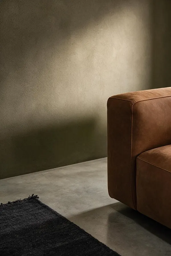 A sparse Brutalist interior with a smooth concrete floor, a textured olive-brown wall, and the corner of a brown leather sofa on the right. A dark shaggy rug sits in the foreground, while soft directional light creates a warm highlight across the wall.
