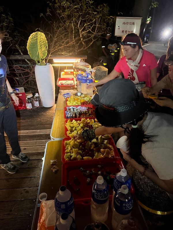 Runners grab refreshments from a well-lit aid station with snacks and drinks during a nighttime trail run race.