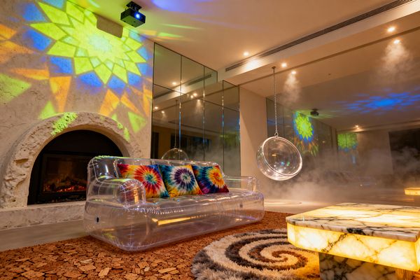 A psychedelic living room with a clear inflatable sofa topped with rainbow tie-dye pillows faces a glowing onyx coffee table on cork flooring and a spiral shag rug. A rough plaster arched fireplace with a live flame sits to the left as kaleidoscopic light patterns wash across mirrored wall panels and the ceiling. A transparent hanging bubble chair on a metal chain hovers in light haze under ceiling spotlights.
