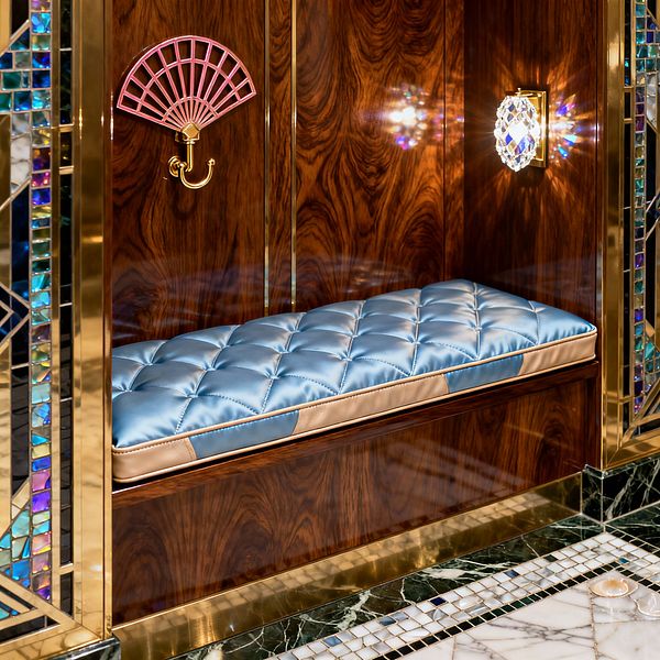 A built-in mudroom niche clad in glossy dark wood with polished brass trim features a powder-blue, diamond‑tufted satin bench cushion finished with champagne piping. A pink fan-shaped brass hook and a faceted crystal wall sconce accent the alcove, while iridescent stained-glass mosaic borders and a floor of dark green marble with white mosaic inlay create an opulent, vintage-inspired setting.