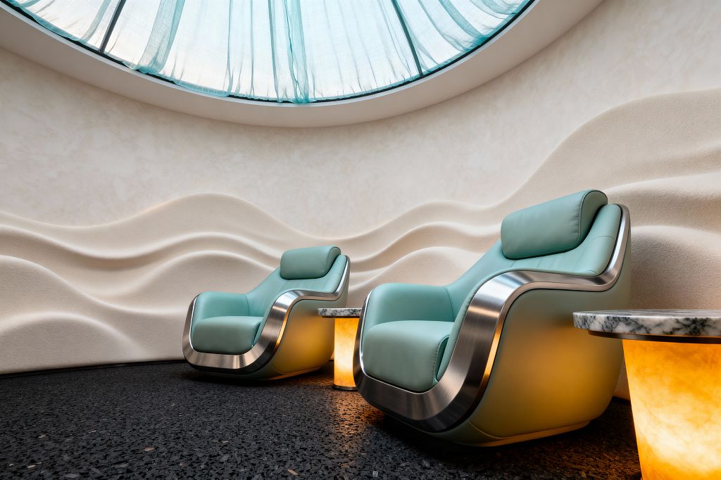 A curved media lounge features two mint leather recliners with brushed stainless-steel frames set against an ivory wall sculpted in flowing wave patterns. A domed skylight with a translucent aqua diffuser bathes the room in soft daylight. Beside each chair, a small table pairs a white marble top with a glowing onyx base, casting warm light onto the dark speckled floor.