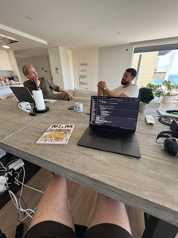 A coworking space scene featuring two individuals engaged in conversation with a laptop and various work items on the table.