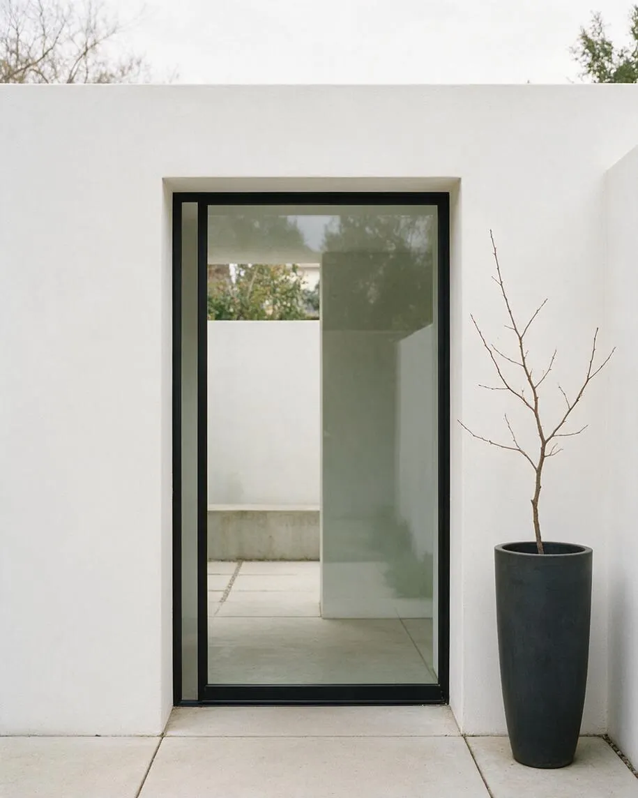 A minimalist exterior with smooth white stucco walls, a tall black-framed glass door, and large pale stone pavers. A slender leafless branch stands in a dark cylindrical planter beside the doorway, creating a calm, sparse composition.
