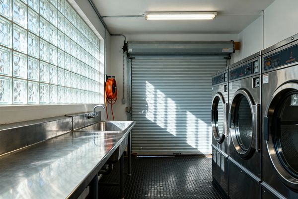 A compact industrial laundry room features a long stainless-steel worktop with a deep sink beneath a translucent glass-block window, while a corrugated roll-up door anchors the far wall. Opposite a row of front-loading stainless washers, the black coin rubber floor, exposed conduit, fluorescent strip light, and an orange hose reel emphasize the utilitarian palette of silver, white, gray, and black lit by crisp daylight.