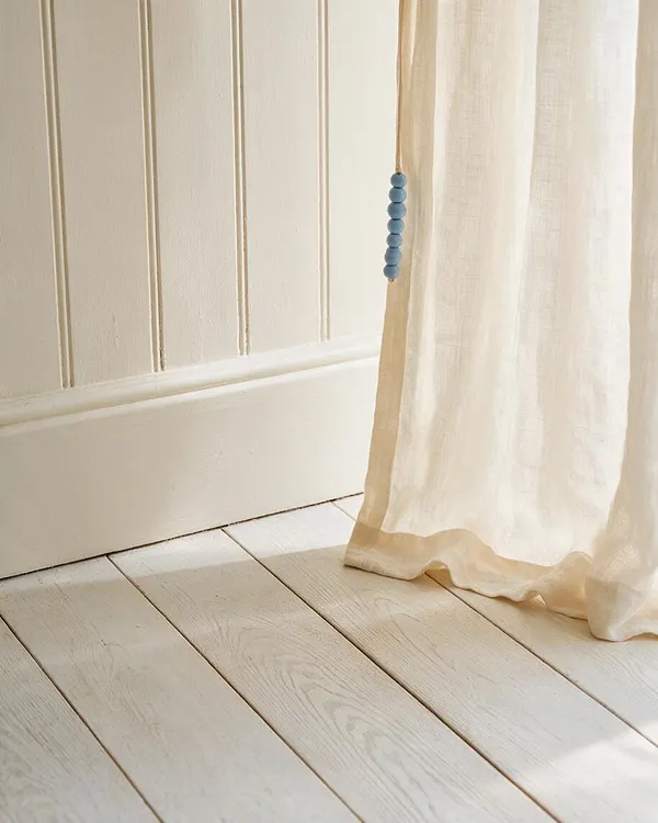 A close-up of a coastal-style interior corner with cream vertical beadboard walls, pale painted floorboards, and a sheer ivory linen curtain trimmed with a soft tan edge. A strand of muted blue beads hangs beside the curtain, adding a subtle seaside accent.
