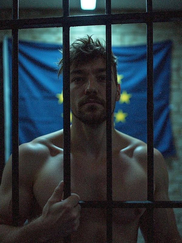 A shirtless man stands behind bars in a dimly lit setting, with a blue flag featuring yellow stars in the background.
