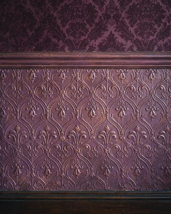 A close-up of a Victorian-style wall treatment in deep plum and mauve tones. The image shows a richly embossed damask-like pattern above dark painted wood trim, creating a layered, ornate surface with a historic feel.