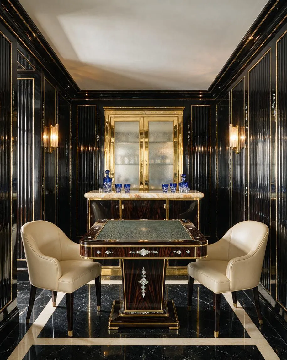 A narrow Art Deco game room with glossy black paneled walls trimmed in gold, a black marble floor with cream border lines, and a central card table. Two cream upholstered chairs flank the table, while a gold-and-glass bar cabinet stands at the back with blue glass decanters and tumblers.