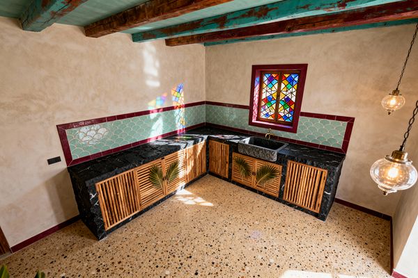 A sunlit Moroccan-style kitchen with an L-shaped layout features a black marble countertop and a dark carved stone sink beneath a jewel-toned stained-glass window. Bamboo slatted cabinet fronts with palm motifs sit below a mint fish-scale zellige backsplash framed in burgundy, while warm beige tadelakt walls, a teal-painted ceiling with rustic wood beams, terrazzo flooring, and glass globe pendants with brass fittings complete the space.