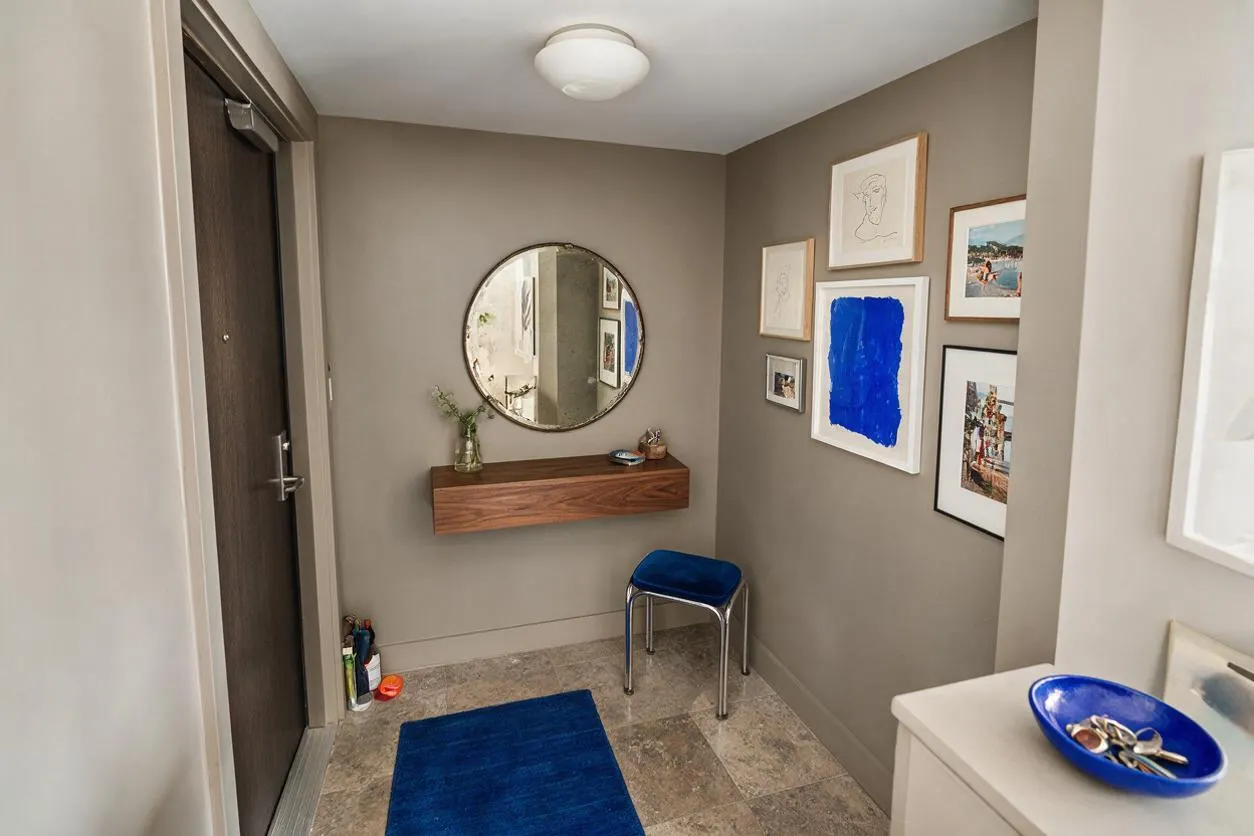 A small eclectic entryway with taupe walls, a dark wood front door, and a round wall mirror above a floating wood console shelf. The space includes a bright blue stool, a blue runner rug, and a gallery wall of mixed framed art, creating a layered, collected look.