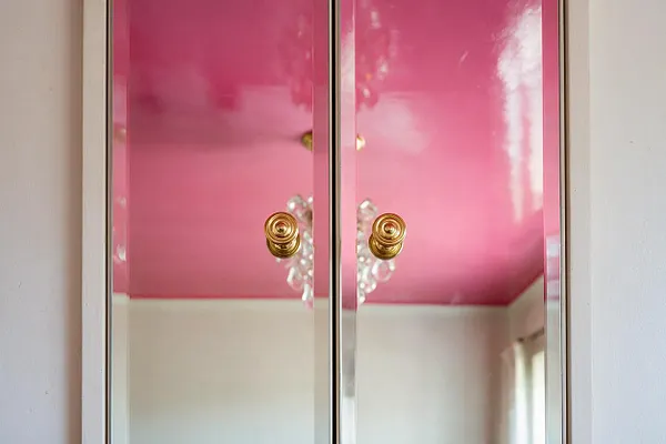 A glossy pink cabinet with mirrored doors fills the frame, each door fitted with a round brass knob. The reflective surface shows a bright room with white walls, a pink ceiling, and a crystal chandelier, creating a glamorous Hollywood Regency look.