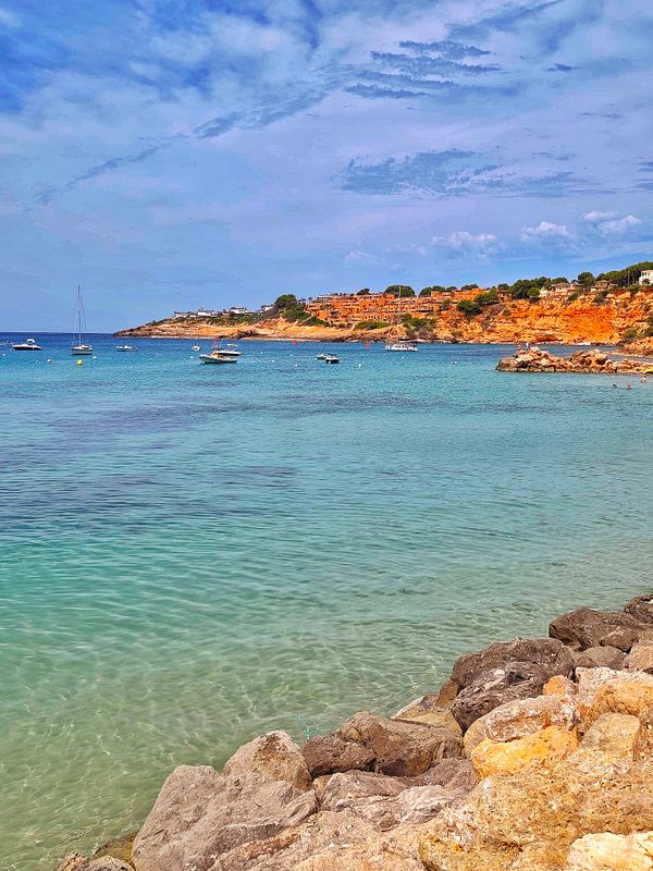 A vibrant beach scene with people swimming and gathering along the shore with blue sky and distant sailboats.
