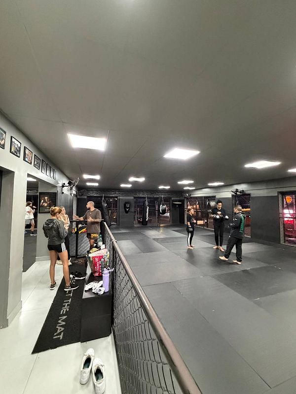 Photograph of an outdoor Muay Thai training session with two fighters sparring inside a ring and a coach instructing.