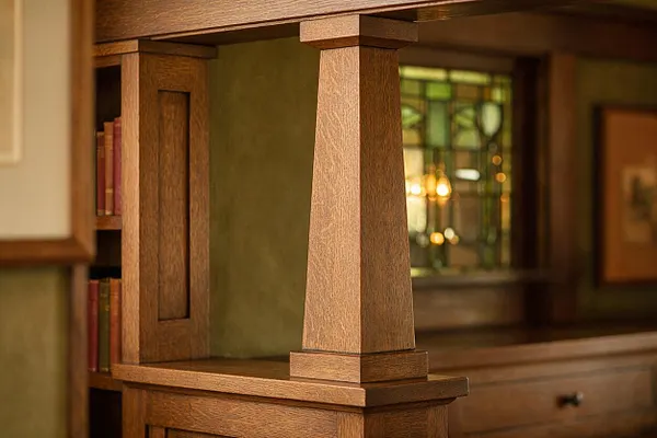 A close-up view of a Craftsman-style built-in featuring a tapered wooden support column with square block details. The surrounding cabinetry is finished in warm stained wood, with books tucked into open shelving and a leaded glass panel softly lit in the background.