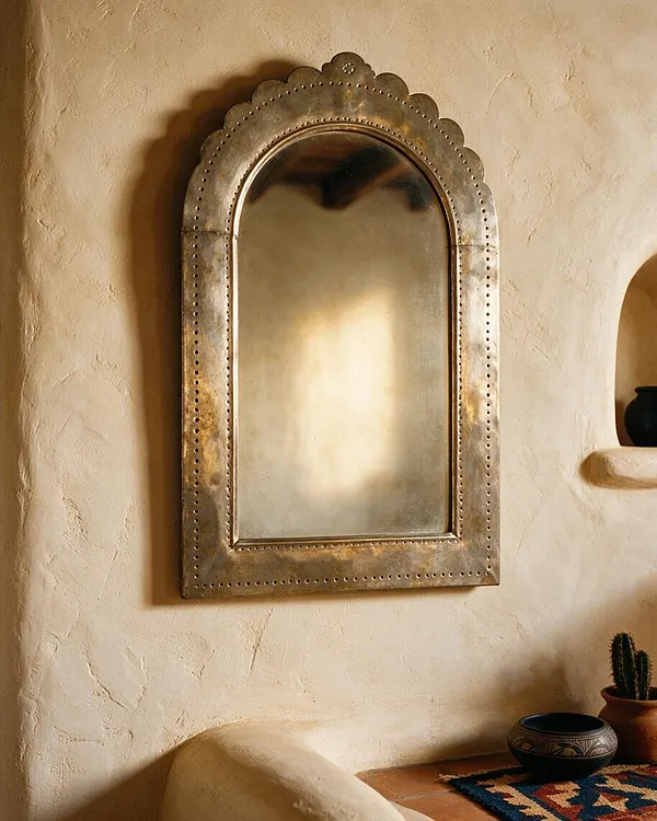 A Southwestern-style wall featuring a tall arched mirror with a scalloped, brass-toned frame and rivet-like detailing mounted on a textured plaster wall. Below it, a curved adobe bench, a woven textile, and small earthenware decor pieces add warmth and handcrafted character.